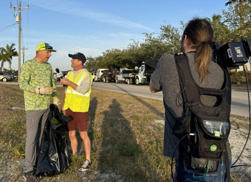 A man holding a trash bag being interviewed by another man in a high visibility vest while they are filmed by a television camera